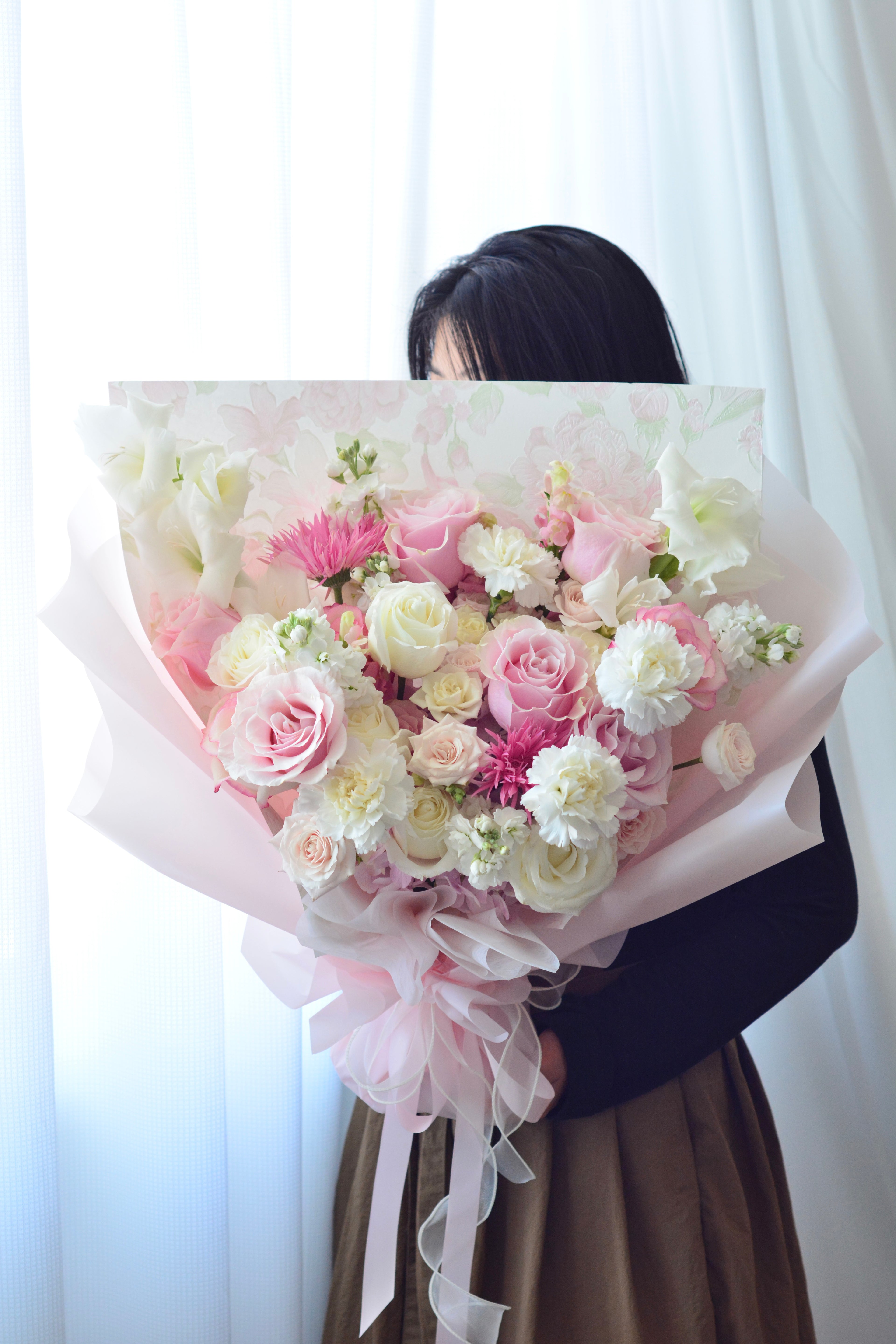 Rose Soufflé bouquet featuring pink and ivory roses, spray roses, carnations, white stocks, and seasonal blooms wrapped in elegant blush paper with luxurious ribbon accents.