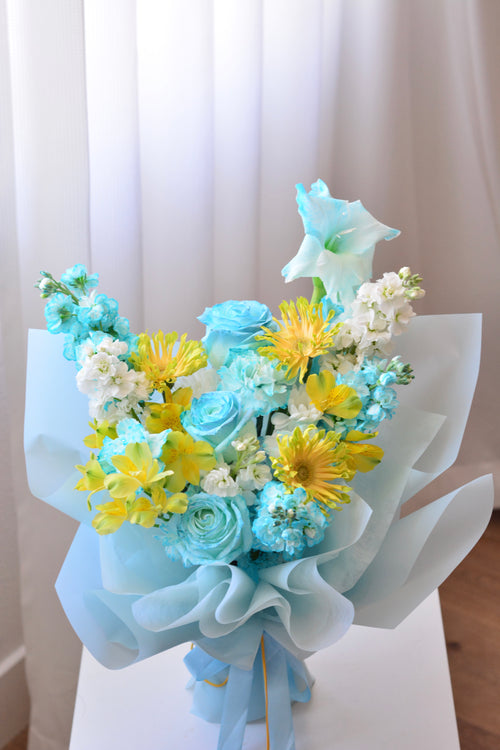 Blue Lemonade bouquet featuring tinted blue roses, yellow gerbera daisies, gladiolus, stock flowers, chrysanthemums, and seasonal blooms wrapped in soft blue paper.