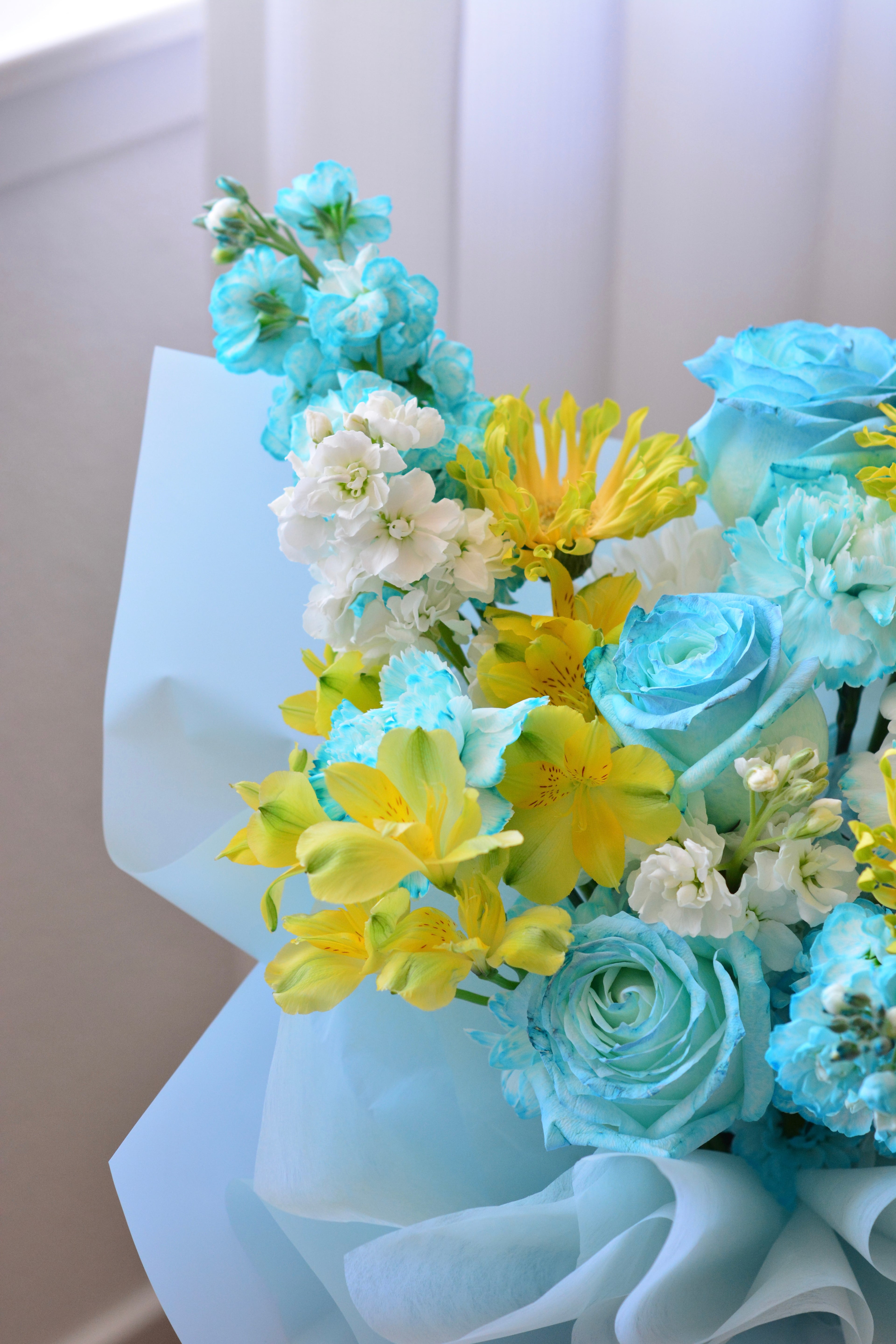 Blue Lemonade bouquet featuring tinted blue roses, yellow gerbera daisies, gladiolus, stock flowers, chrysanthemums, and seasonal blooms wrapped in soft blue paper.