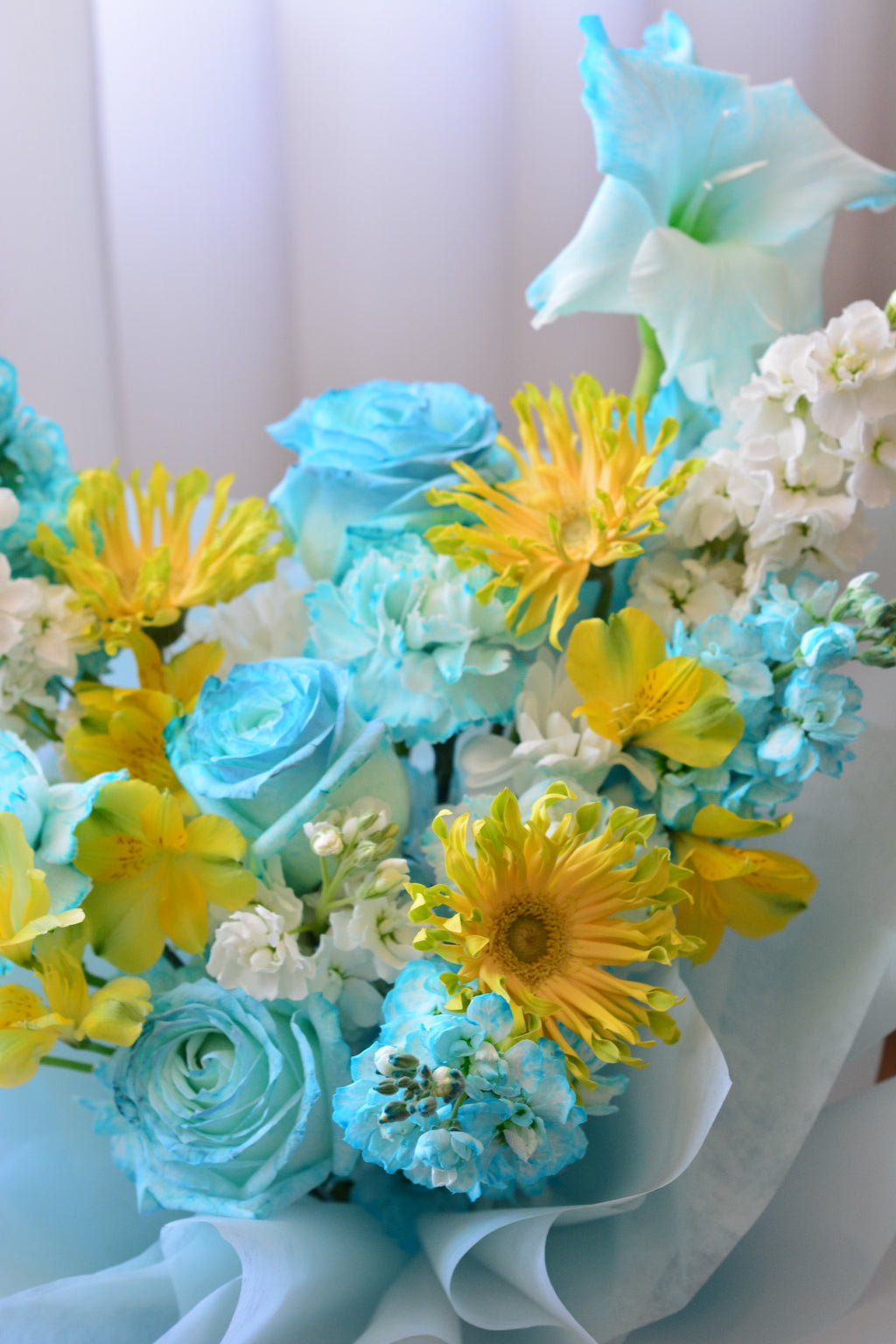 Blue Lemonade bouquet featuring tinted blue roses, yellow gerbera daisies, gladiolus, stock flowers, chrysanthemums, and seasonal blooms wrapped in soft blue paper.