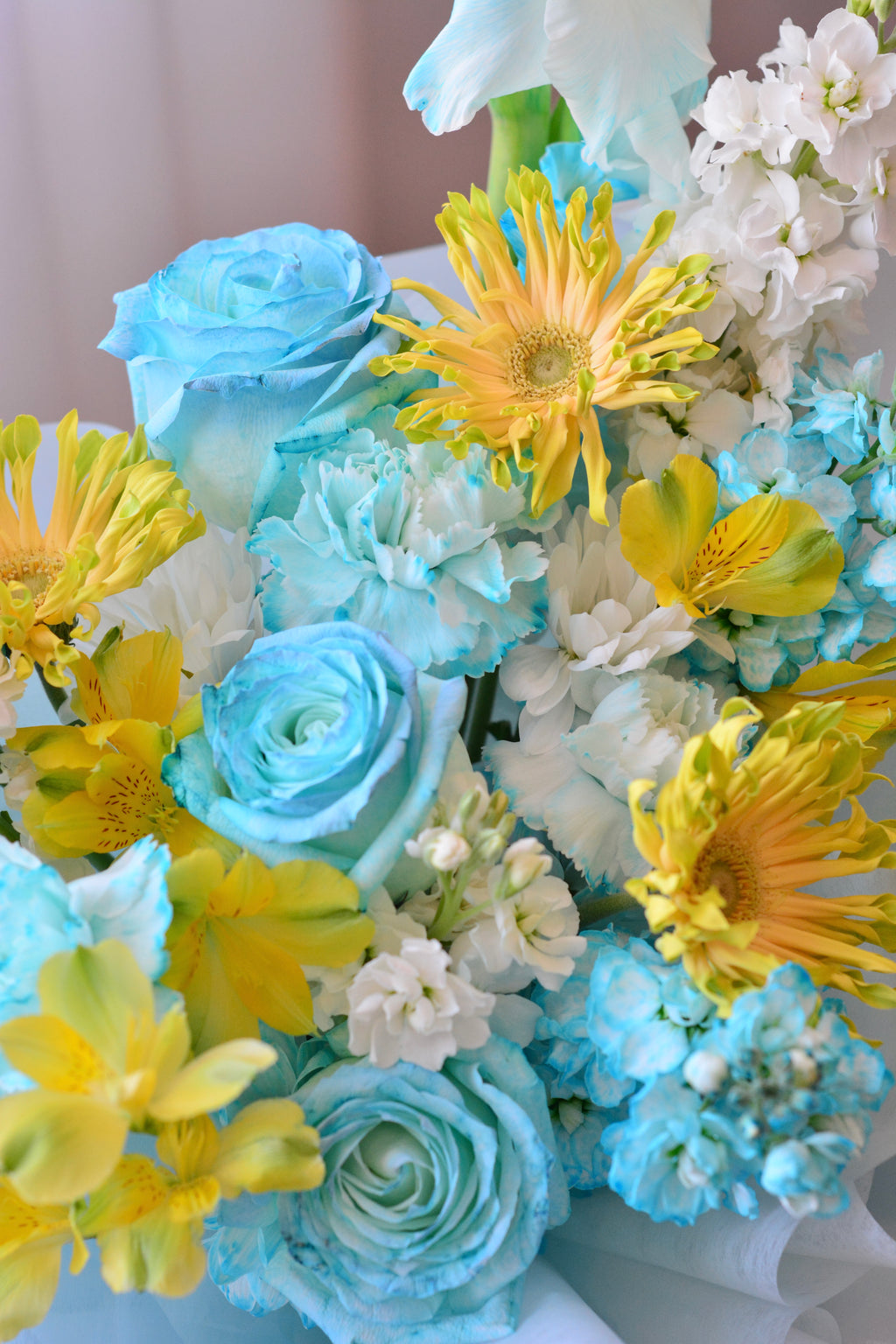 Blue Lemonade bouquet featuring tinted blue roses, yellow gerbera daisies, gladiolus, stock flowers, chrysanthemums, and seasonal blooms wrapped in soft blue paper.
