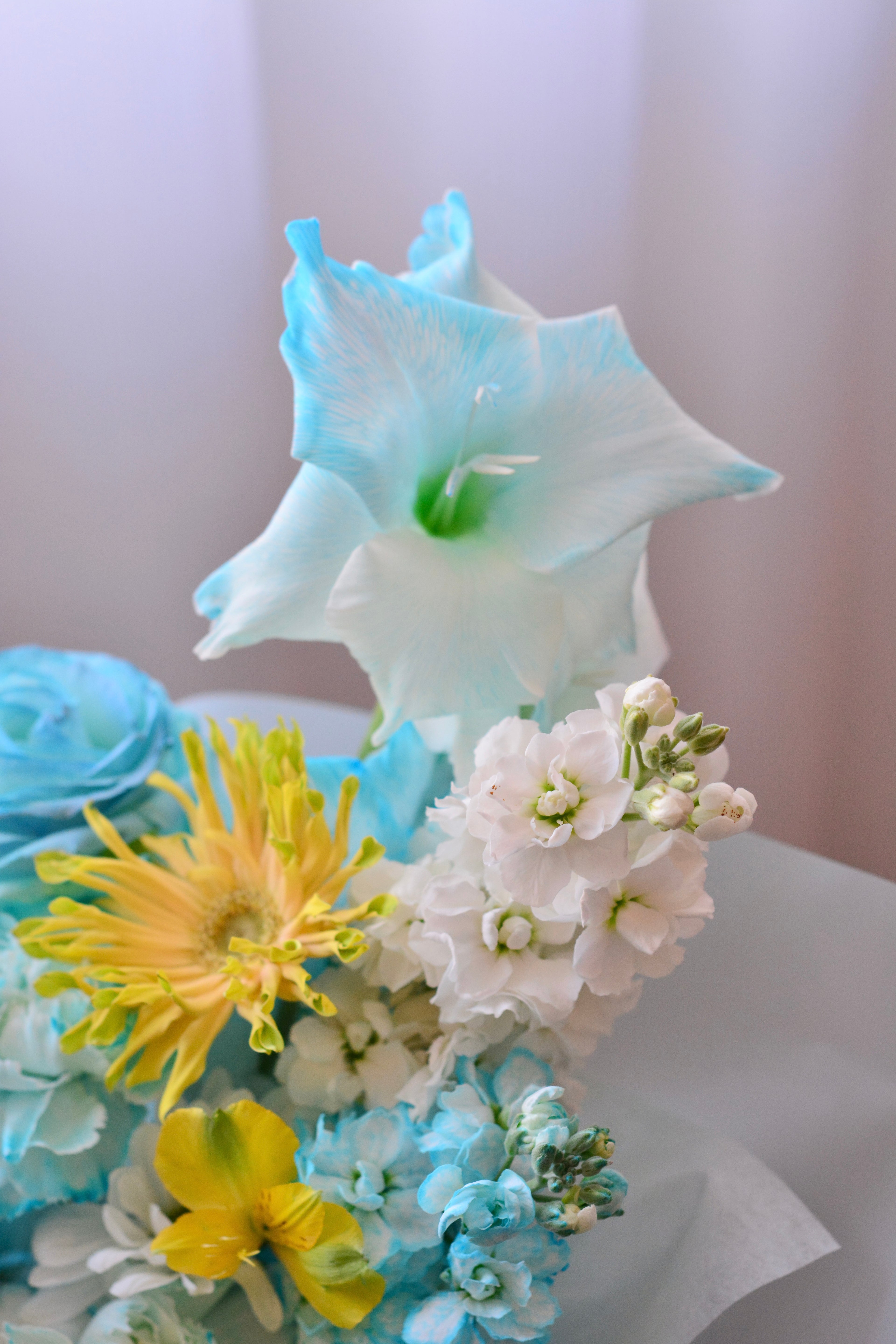 Blue Lemonade bouquet featuring tinted blue roses, yellow gerbera daisies, gladiolus, stock flowers, chrysanthemums, and seasonal blooms wrapped in soft blue paper.