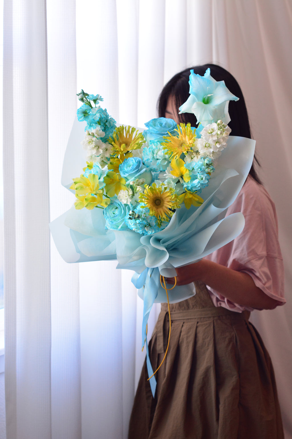 Blue Lemonade bouquet featuring tinted blue roses, yellow gerbera daisies, gladiolus, stock flowers, chrysanthemums, and seasonal blooms wrapped in soft blue paper.