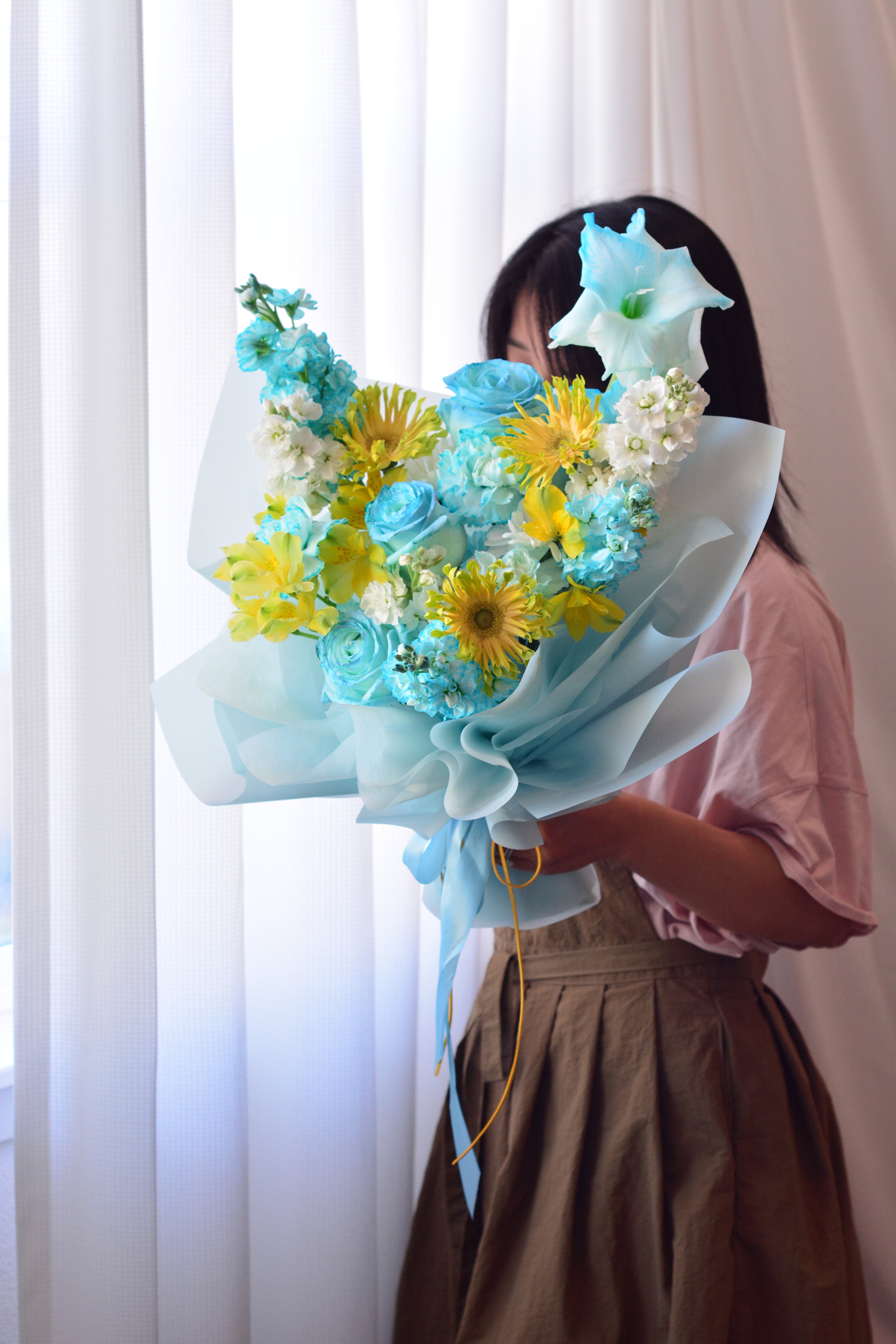 Blue Lemonade bouquet featuring tinted blue roses, yellow gerbera daisies, gladiolus, stock flowers, chrysanthemums, and seasonal blooms wrapped in soft blue paper.